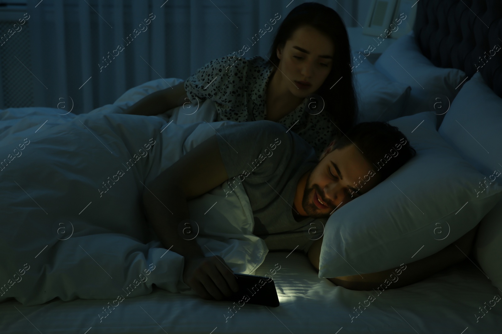 Photo of Distrustful young woman peering into boyfriend's smartphone in bed at night