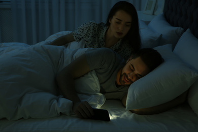 Photo of Distrustful young woman peering into boyfriend's smartphone in bed at night