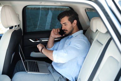 Photo of Attractive young man working with laptop and talking on phone in luxury car