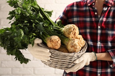 Woman holding wicker basket with raw celery roots near white brick wall, closeup