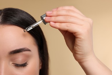 Photo of Young woman applying essential oil onto face on beige background, closeup