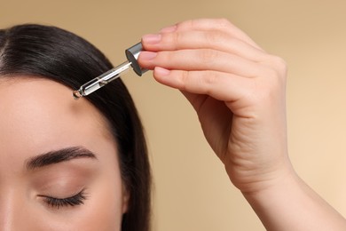 Young woman applying essential oil onto face on beige background, closeup