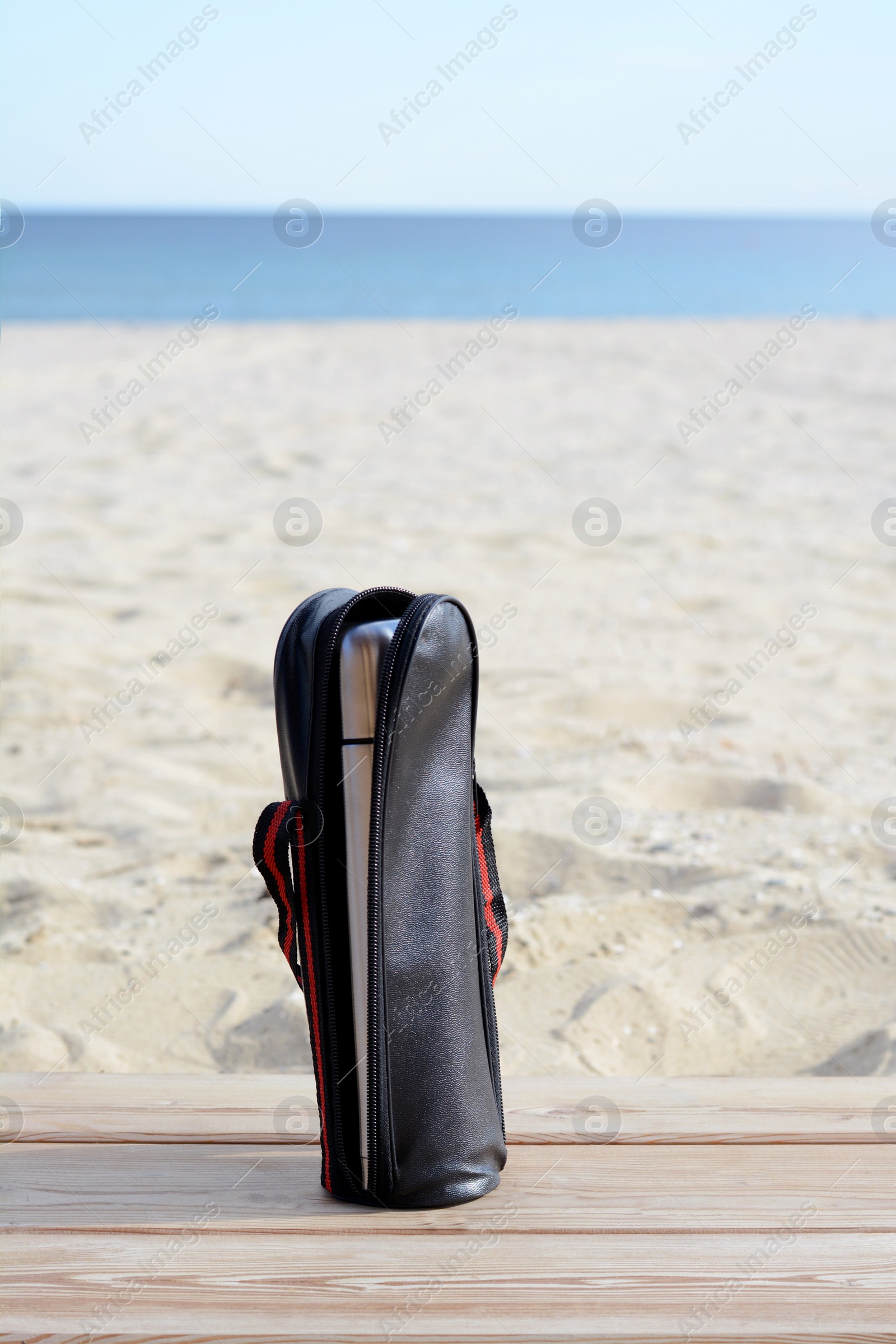 Photo of Metallic thermos in black stylish case on wooden surface near sea, space for text