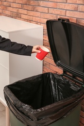 Photo of Young woman throwing coffee cup in trash bin indoors, closeup. Waste recycling