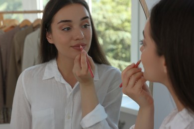 Photo of Beautiful young woman applying cosmetic pencil on lips near mirror indoors