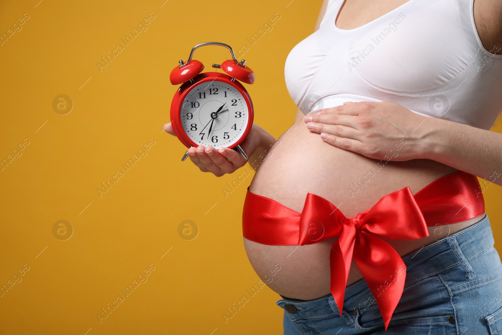 Photo of Young pregnant woman with alarm clock and red bow on yellow background, closeup. Time to give birth