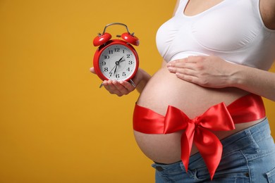 Photo of Young pregnant woman with alarm clock and red bow on yellow background, closeup. Time to give birth