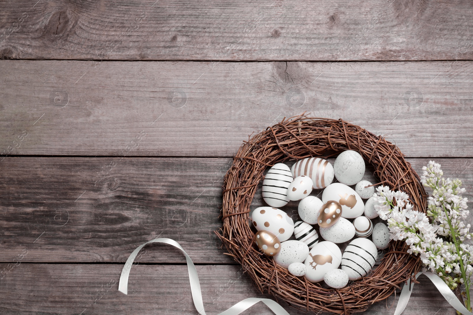 Photo of Many painted Easter eggs, branch of lilac flowers and ribbon on wooden table, flat lay. Space for text