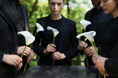 People with calla lily flowers near granite tombstone at cemetery outdoors, closeup. Funeral ceremony