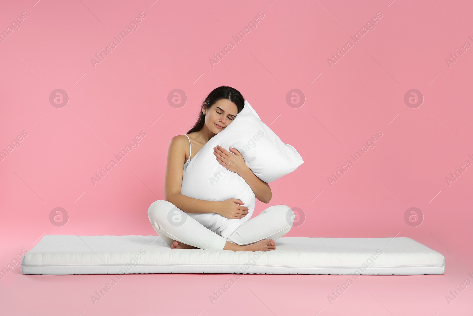 Photo of Young woman sitting on soft mattress and holding pillow against pink background