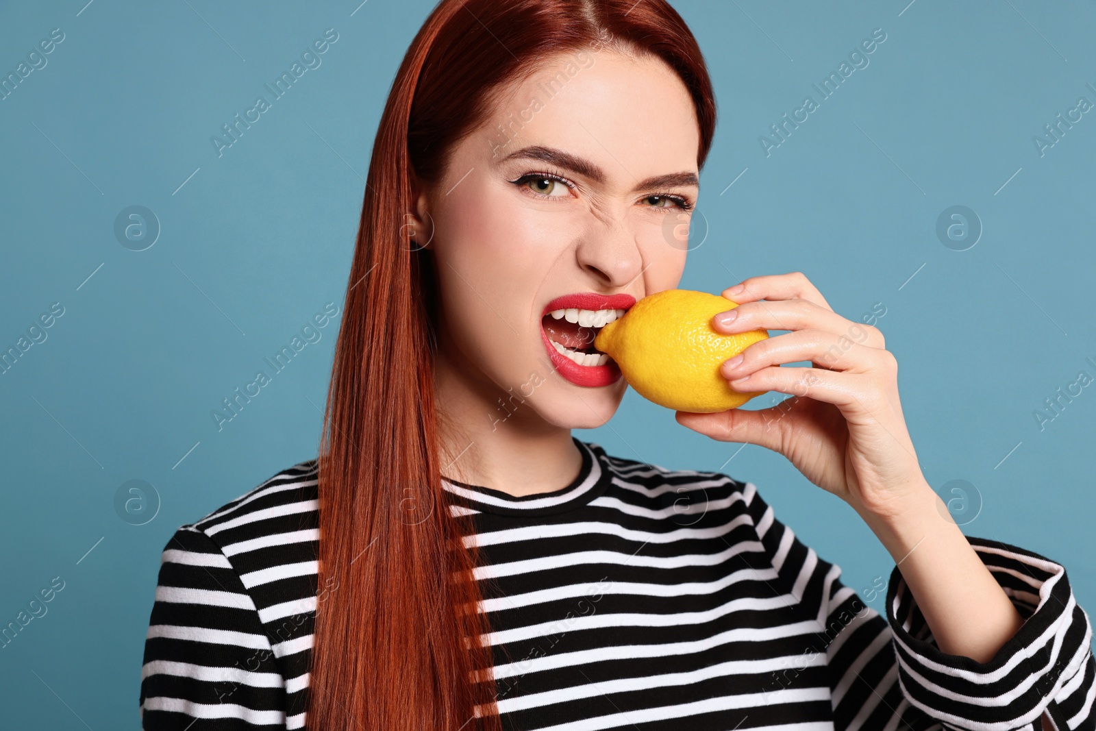 Photo of Woman with red dyed hair biting whole lemon on light blue background