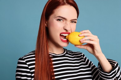 Woman with red dyed hair biting whole lemon on light blue background