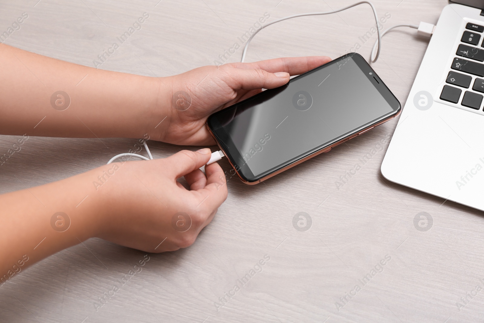 Photo of Woman connecting smartphone with charge cable to laptop on light grey table, closeup. Space for text