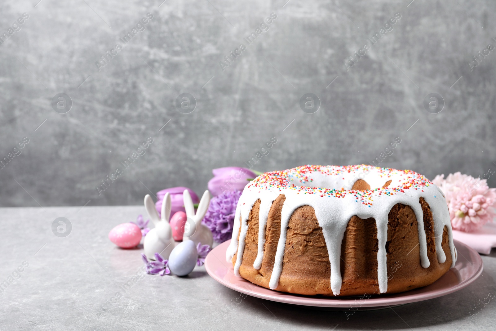 Photo of Glazed Easter cake with sprinkles, painted eggs and flowers on grey table, space for text