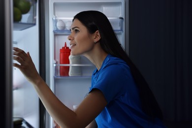 Young smiling woman looking into modern refrigerator at night