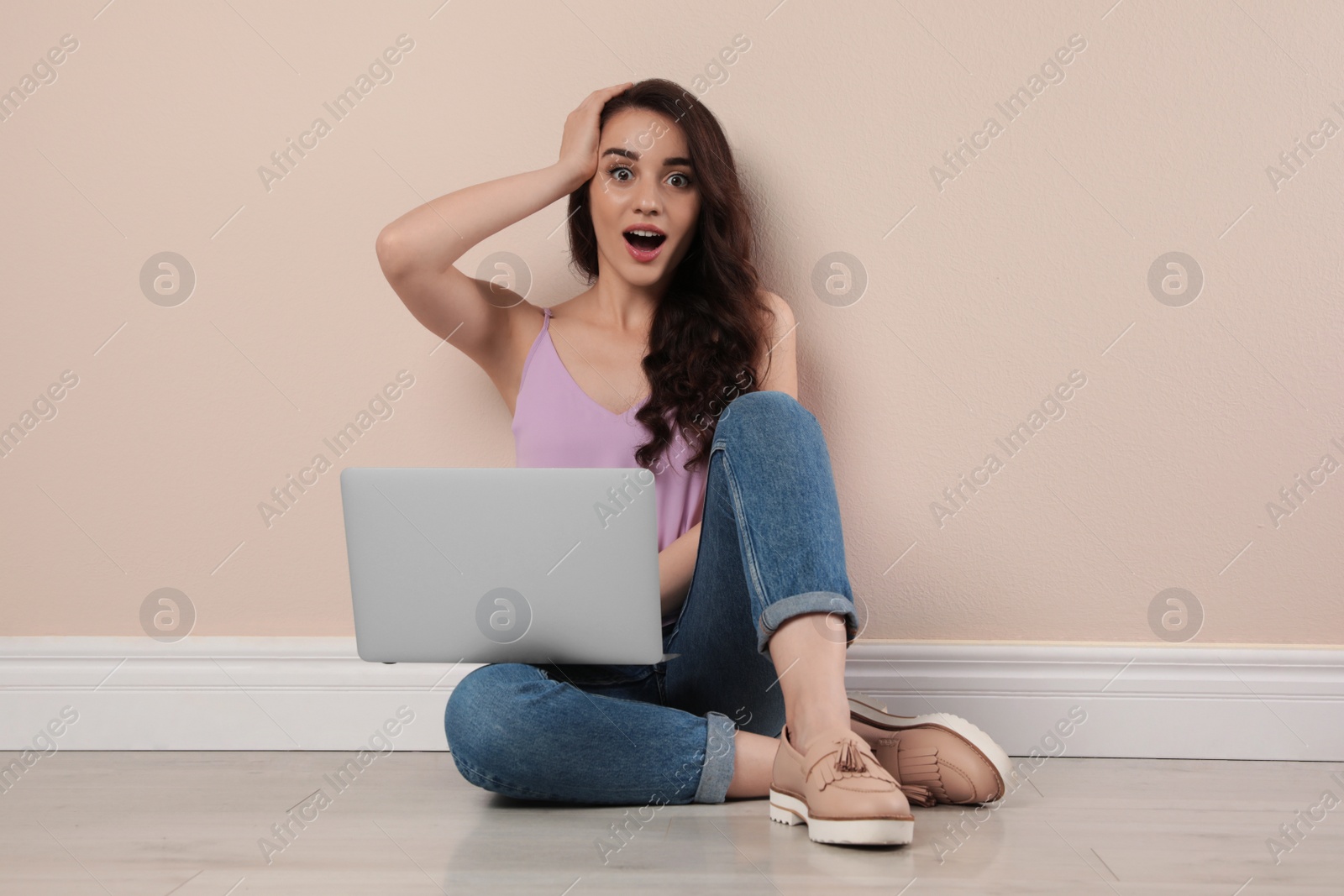 Photo of Shocked young woman sitting with laptop near beige wall