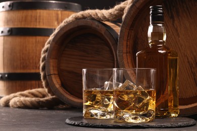 Photo of Whiskey with ice cubes in glasses, bottle and wooden barrels on black table, space for text