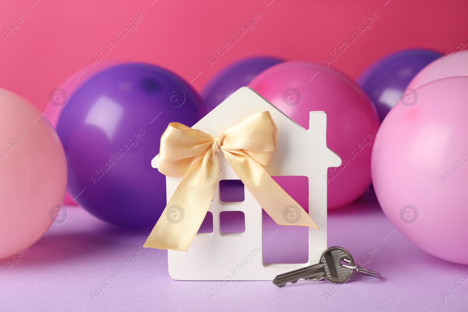 Photo of House model with bow, key and colorful balloons on violet table. Housewarming party