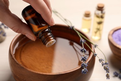 Woman dripping lavender essential oil from bottle into bowl at white table, closeup