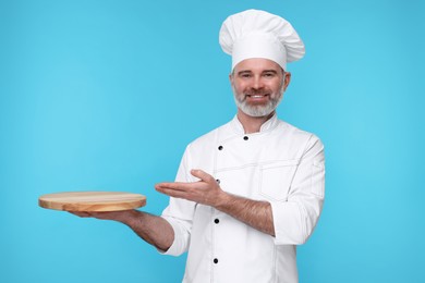 Photo of Happy chef in uniform showing wooden board on light blue background