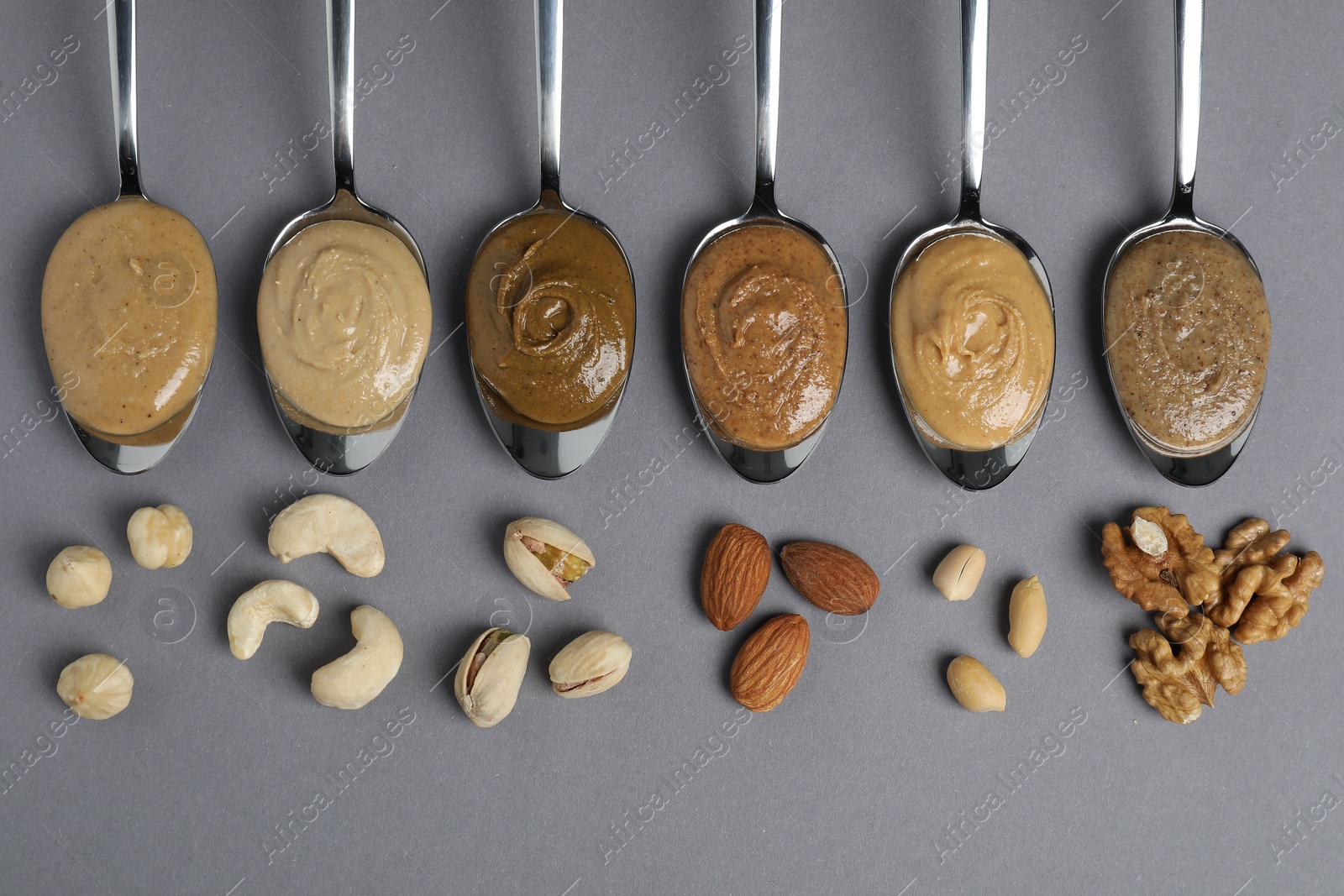 Photo of Tasty nut butters in spoons and raw nuts on gray table, flat lay