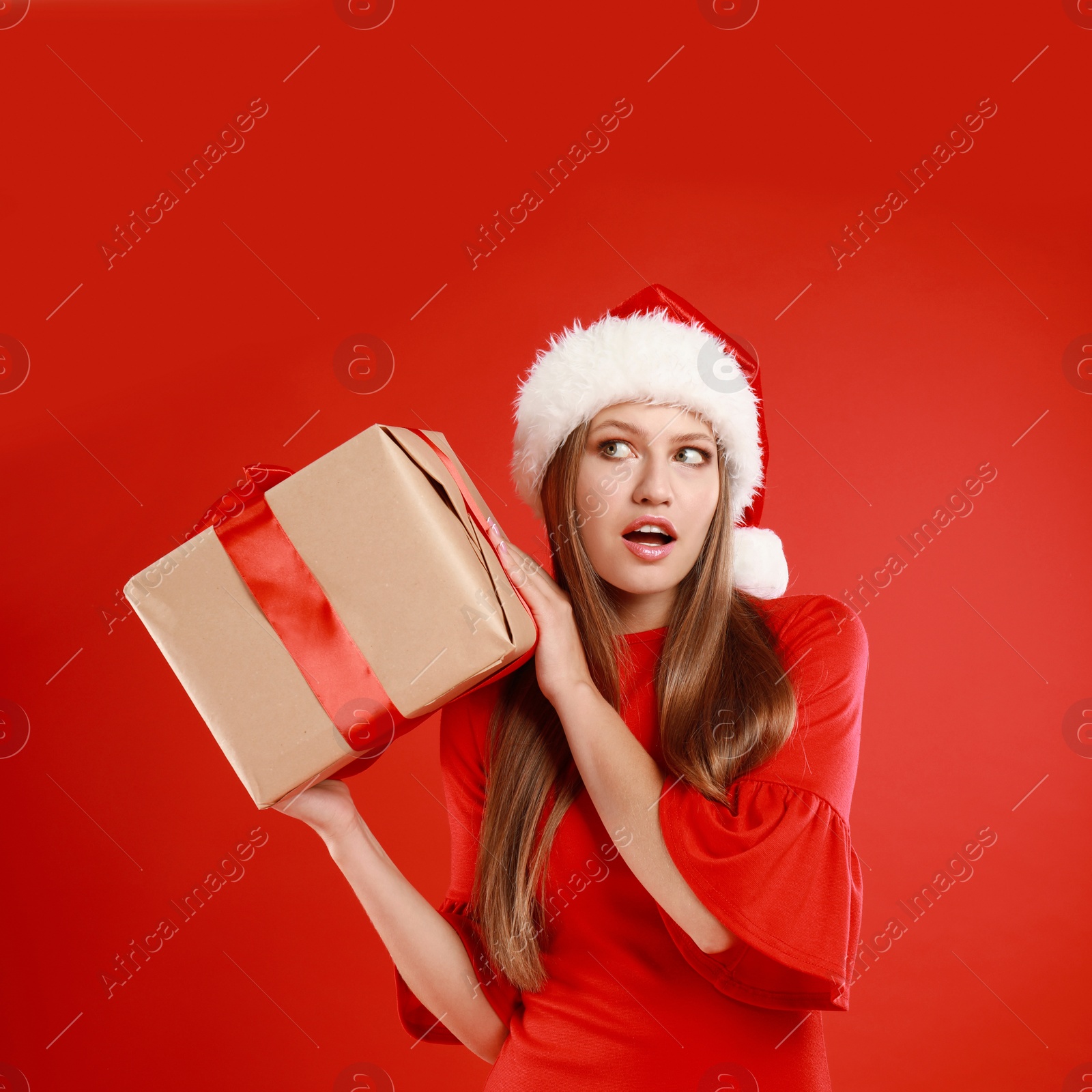 Photo of Emotional young woman in Santa hat with Christmas gift on red background