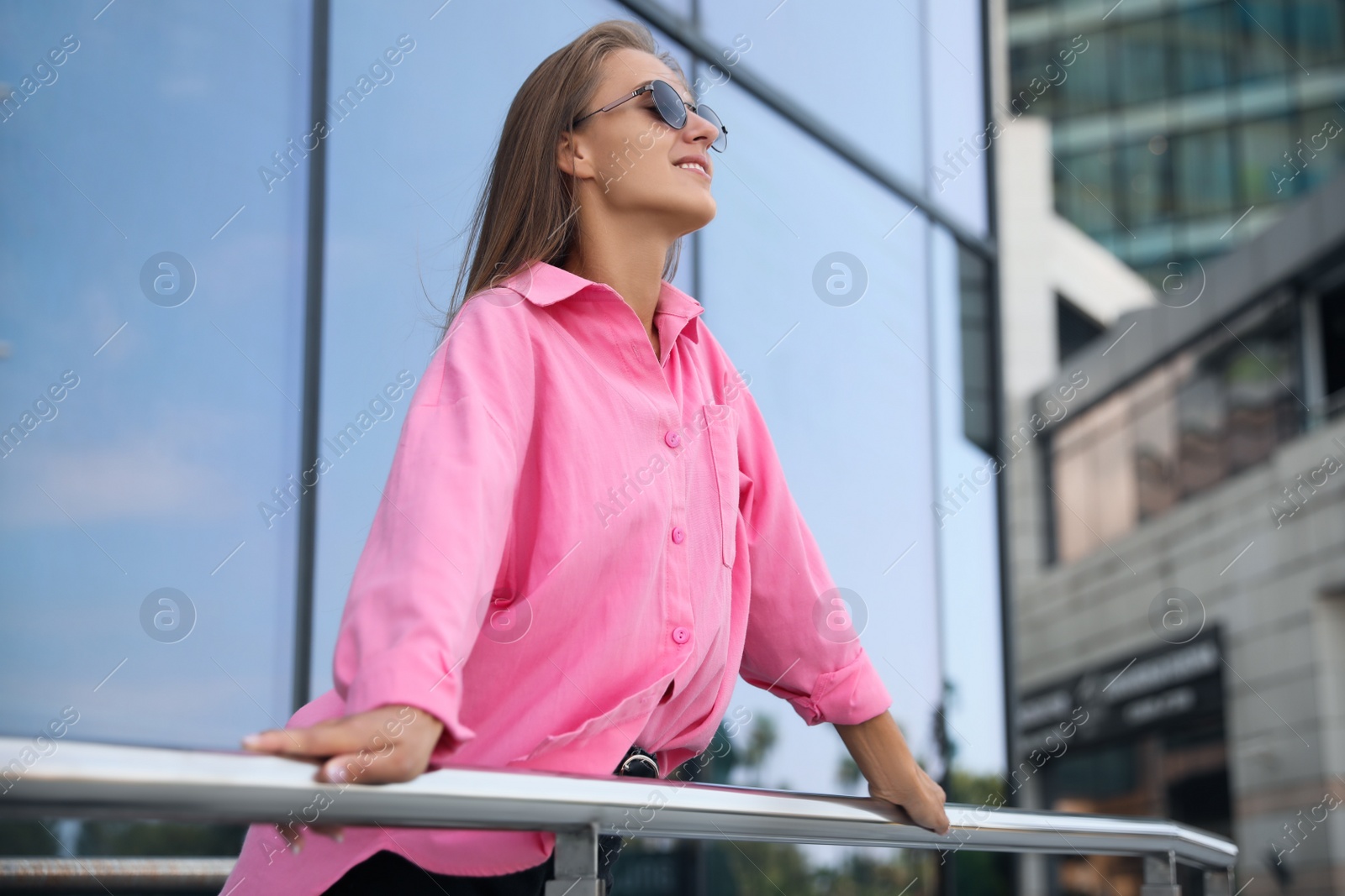 Photo of Beautiful young woman in stylish sunglasses holding onto railing near building outdoors