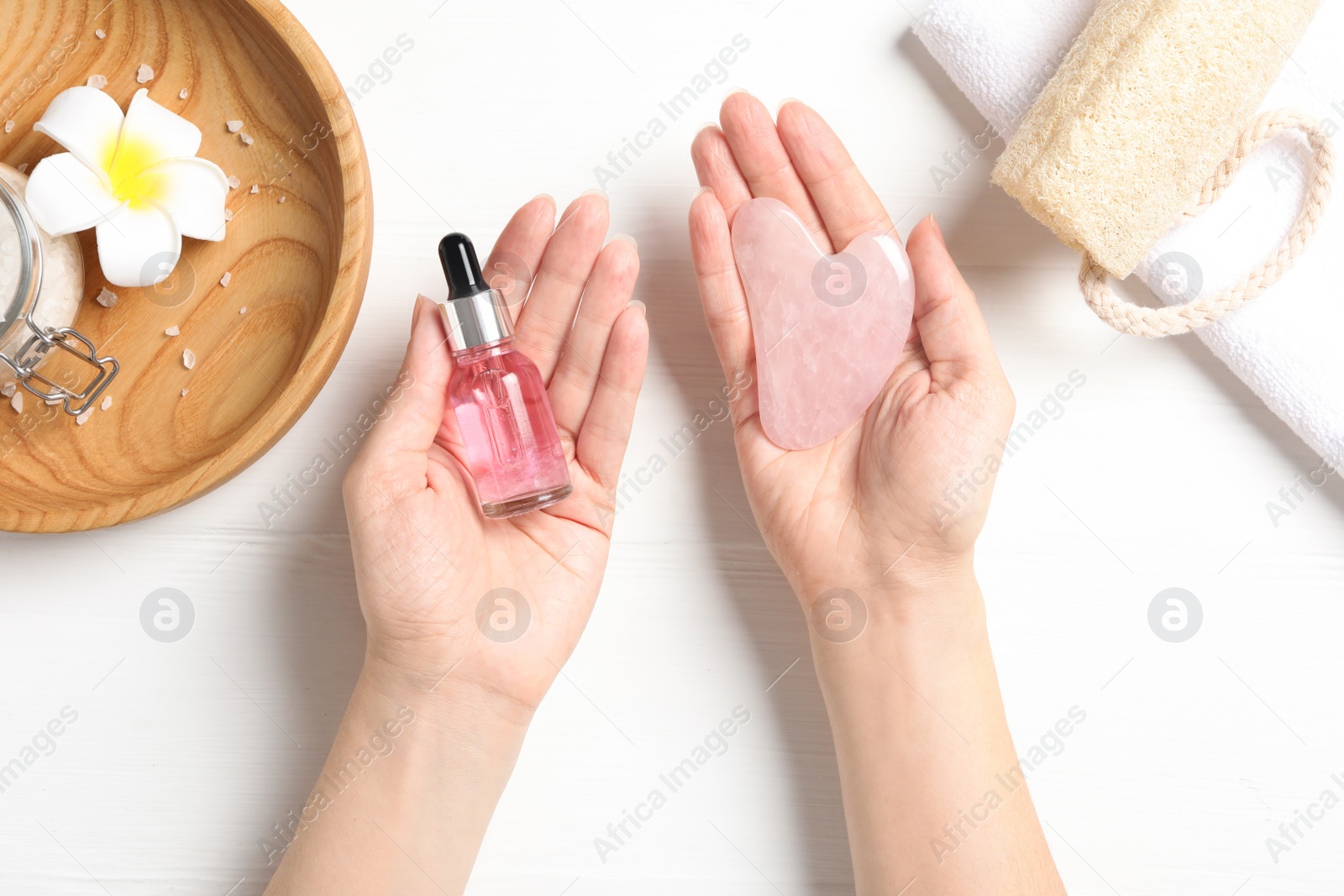 Photo of Woman holding gua sha tool and cosmetic over white wooden table, top view