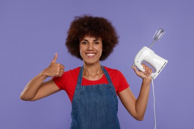 Photo of Happy young woman in apron holding mixer and showing thumb up on purple background