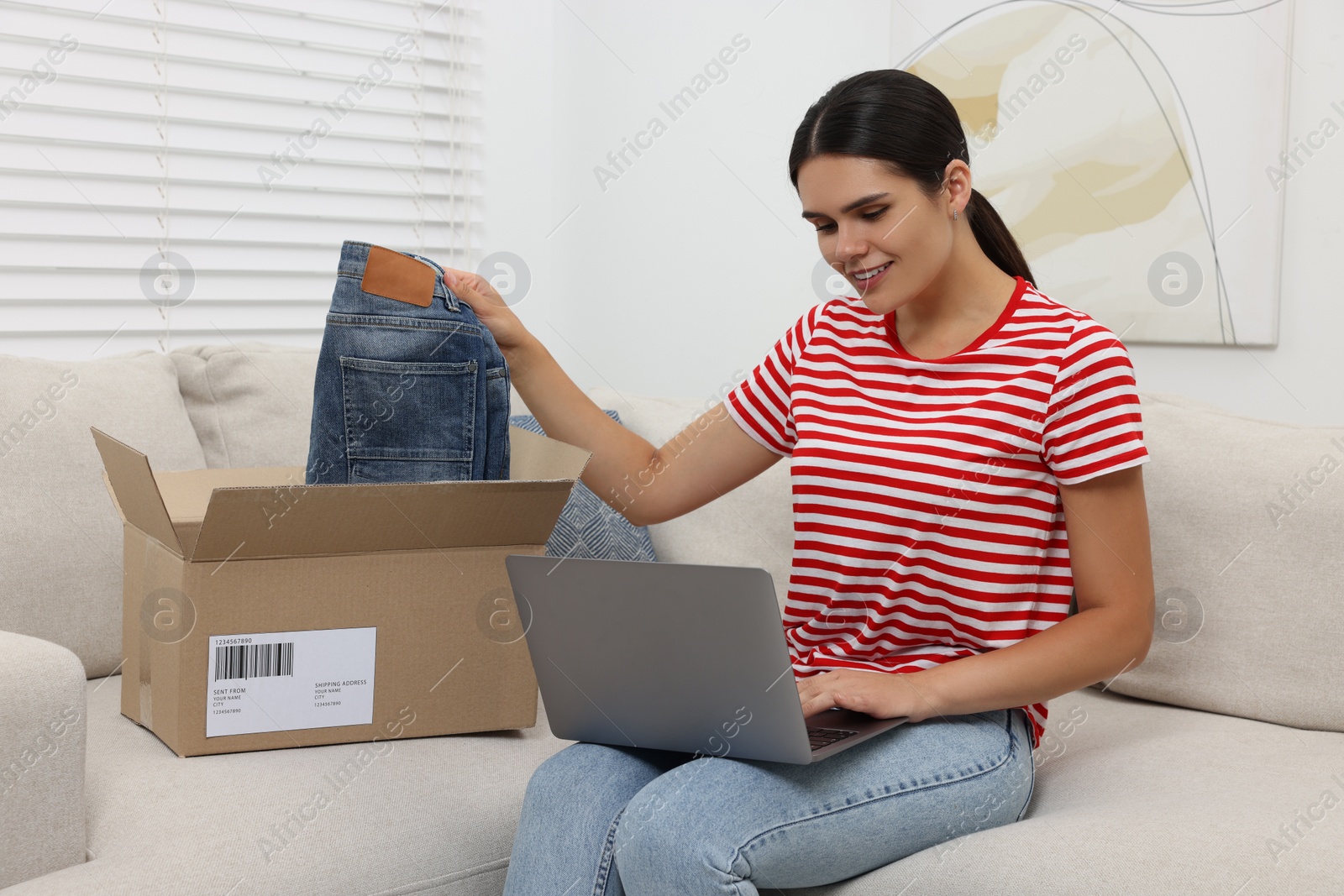 Photo of Young woman with just unpacked new jeans using laptop on sofa at home. Online shopping