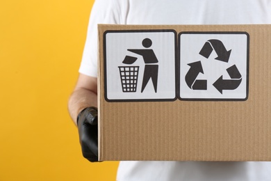 Photo of Courier holding cardboard box with different packaging symbols on yellow background, closeup. Parcel delivery