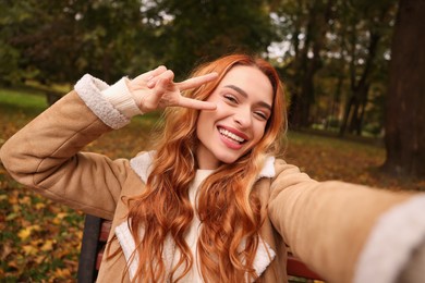 Photo of Portrait of happy woman taking selfie and showing peace sign in autumn park