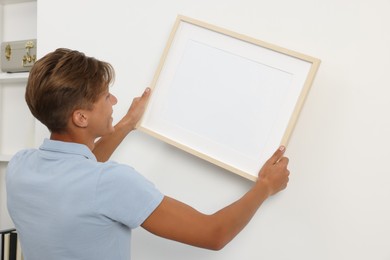 Photo of Young man hanging picture frame on white wall indoors, back view