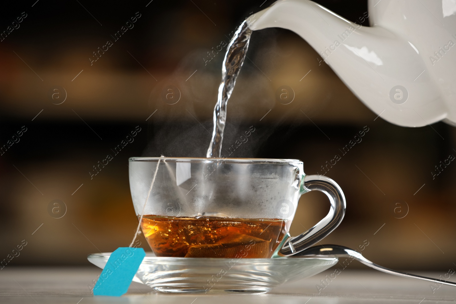 Photo of Pouring hot water into glass cup with tea bag on table against blurred background