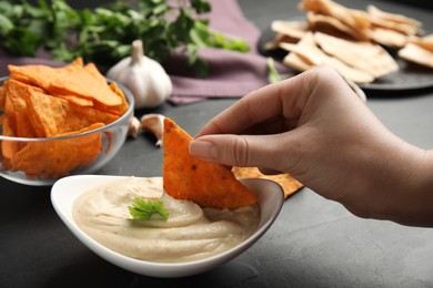 Woman dipping nacho chip into hummus at grey table, closeup