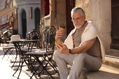 Photo of Handsome senior man sitting on doorstep, using smartphone and drinking coffee outdoors, space for text