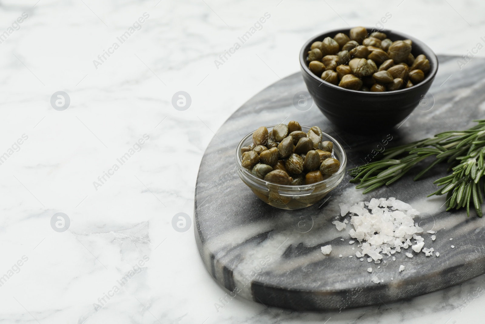 Photo of Delicious pickled capers, salt and rosemary twigs on white marble table, space for text