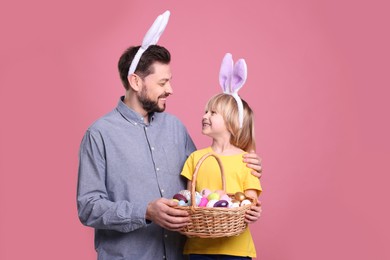 Father and son in bunny ears headbands with wicker basket of painted Easter eggs on pink background. Space for text