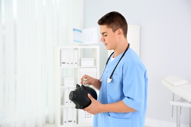Photo of Young medical assistant putting money into piggy bank in clinic. Space for text