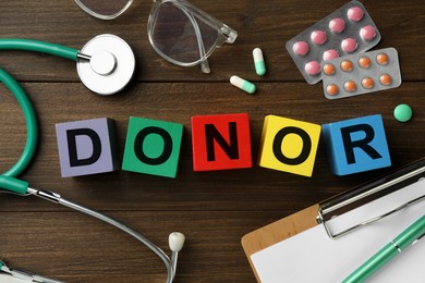 Photo of Word Donor made of colorful cubes, pills, clipboard and stethoscope on wooden table, flat lay