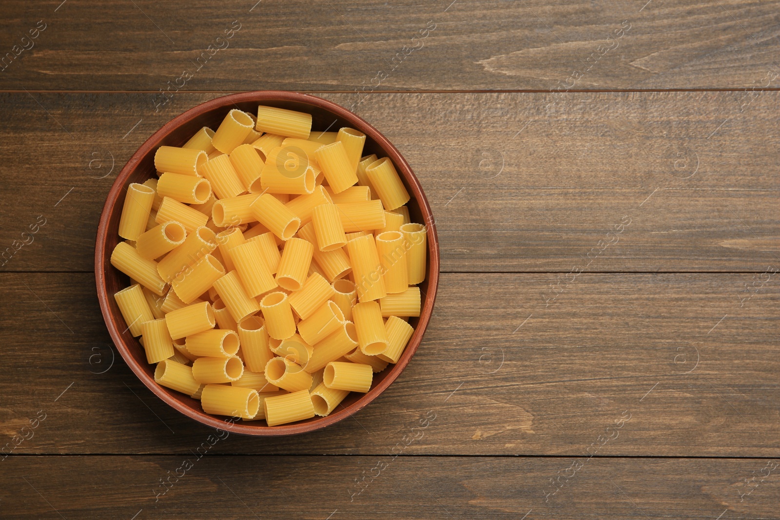 Photo of Raw rigatoni pasta in bowl on wooden table, top view. Space for text