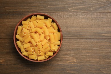 Photo of Raw rigatoni pasta in bowl on wooden table, top view. Space for text