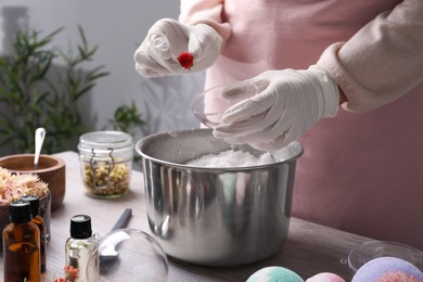 Photo of Woman in gloves putting dried flower into bath bomb mold at wooden table indoors, closeup