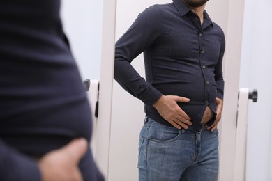 Man wearing tight shirt in front of mirror indoors, closeup. Overweight problem