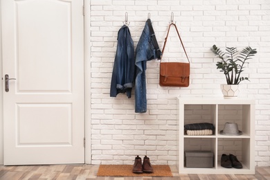 Stylish hallway interior with door, comfortable furniture and clothes on brick wall