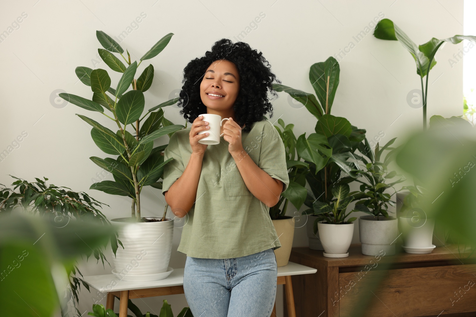 Photo of Relaxing atmosphere. Happy woman with cup of hot drink near beautiful houseplants in room