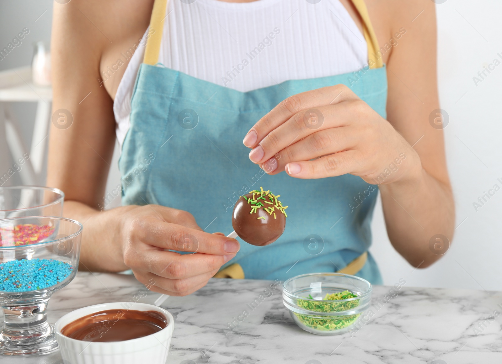 Photo of Young woman decorating tasty cake pop with green sprinkles at white marble table, closeup