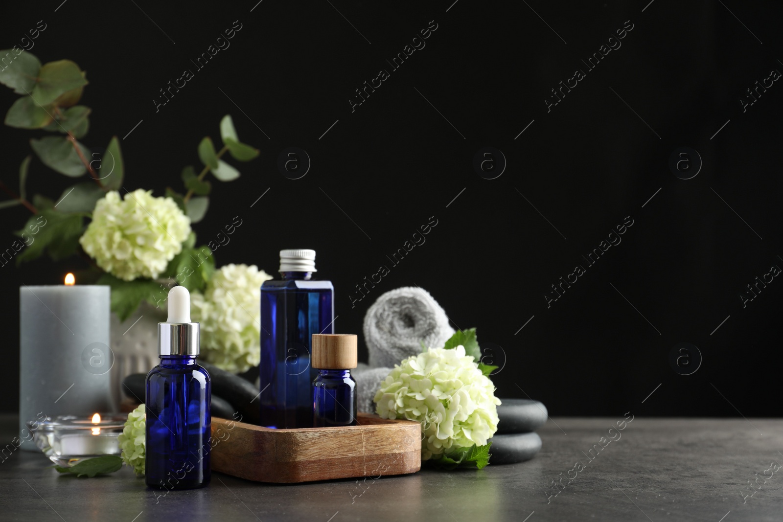 Photo of Spa composition. Cosmetic products, burning candles and hydrangea flowers on gray table against black background, space for text