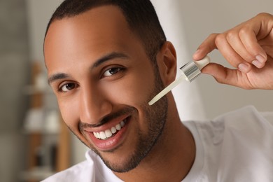 Photo of Handsome man applying cosmetic serum onto face in bathroom, closeup