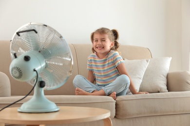 Photo of Little girl enjoying air flow from fan on sofa in living room. Summer heat